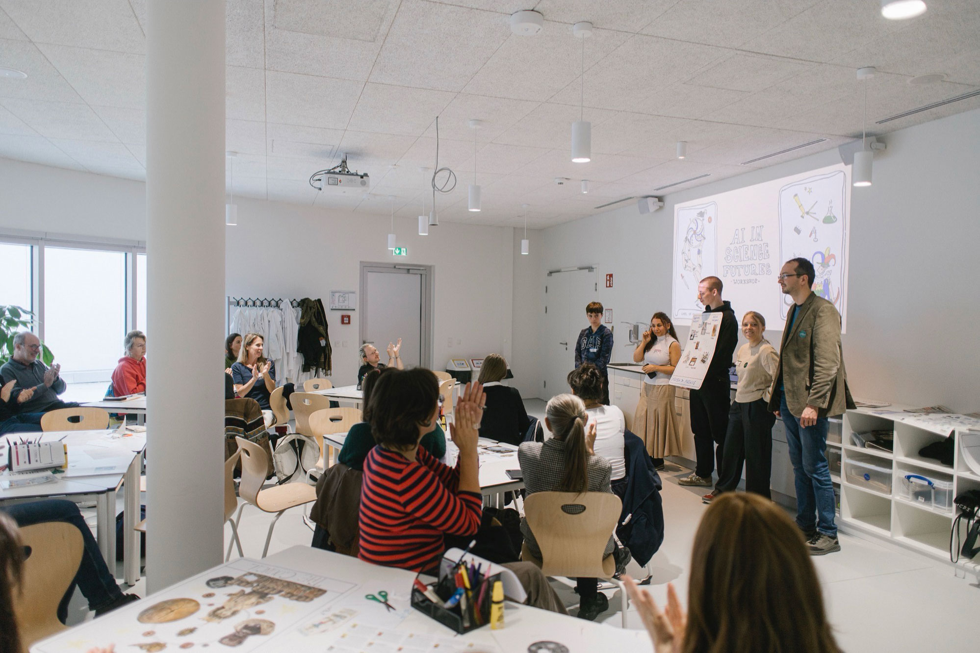 Photo from the workshop, people sitting around tables with craft supplies, applauding. A group of people presenting a poster in front of a projection of the workshop title.